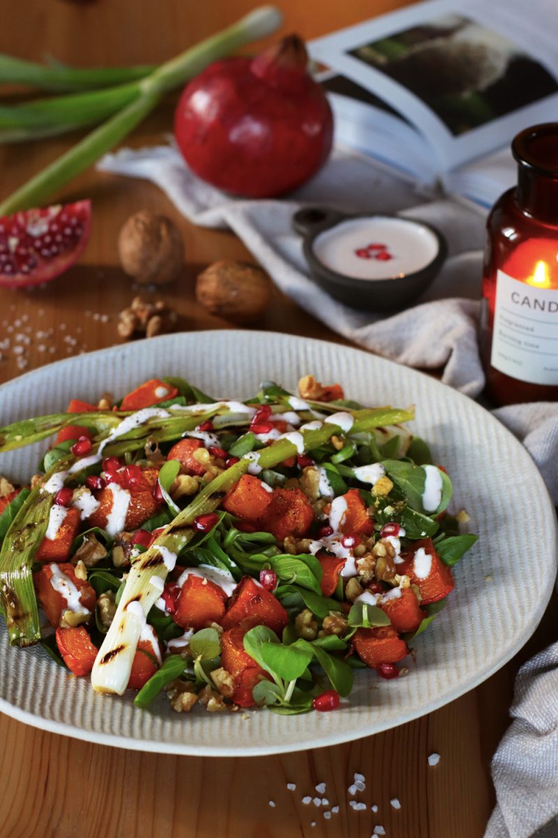 Insalata tiepida di zucca arrostita con cipollotto grigliato, noci, melagrana e salsa di yogurt, disposta su un piatto bianco. Sullo sfondo, ingredienti freschi e un libro di ricette.