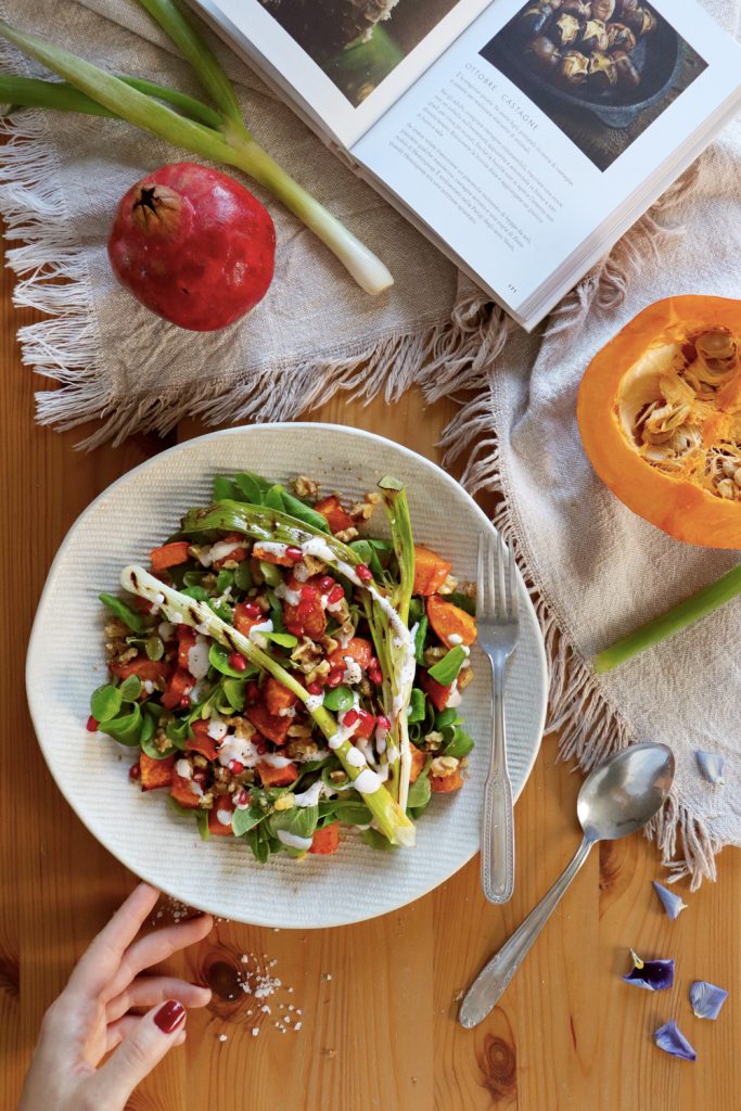Insalata tiepida di zucca arrostita con cipollotto grigliato, noci, melagrana e salsa di yogurt, disposta su un piatto bianco. Sullo sfondo, ingredienti freschi e un libro di ricette.