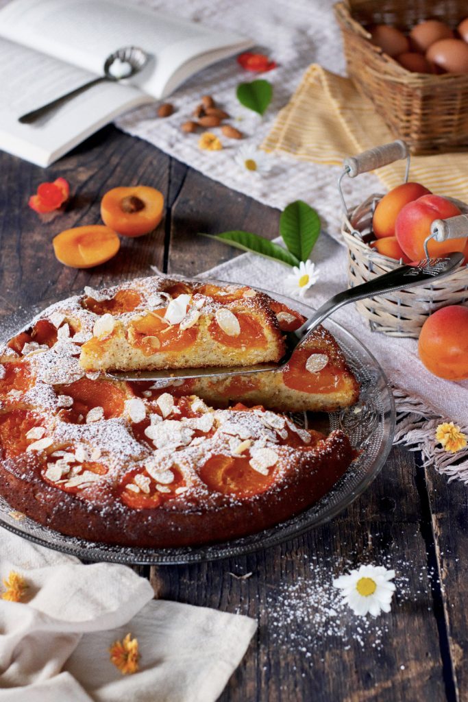 Torta soffice alla ricotta con albicocche fresche e mandorle a lamelle, servita su piatto di vetro con spolverata di zucchero a velo, su tavolo rustico in legno con frutta e libro aperto sullo sfondo.