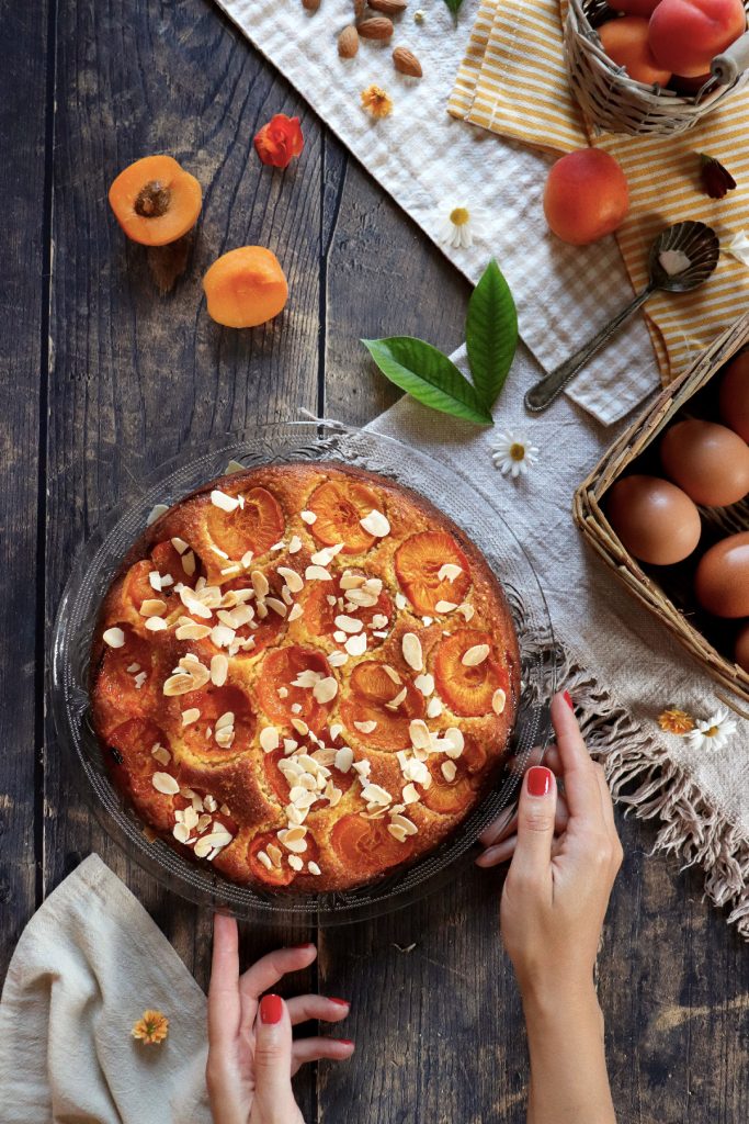 Mani femminili con smalto rosso che servono una torta alla ricotta con albicocche e mandorle, vista dall’alto su tavolo rustico con ingredienti freschi, uova e frutta di stagione.
