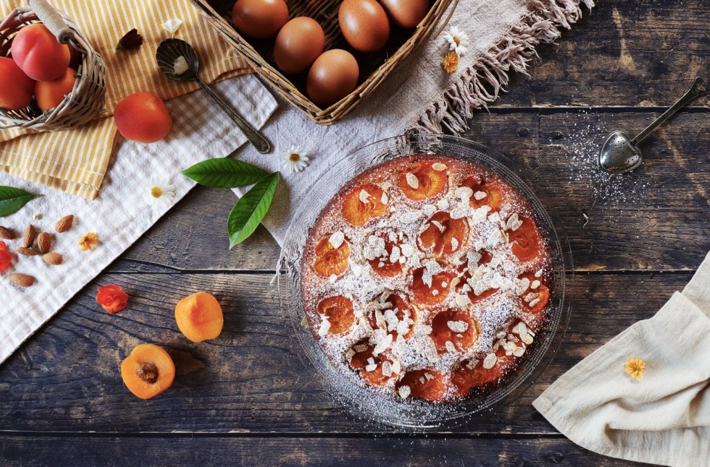 Vista dall’alto di torta alla ricotta con albicocche e mandorle in teglia di vetro, decorata con zucchero a velo, su tavolo rustico con uova, frutta fresca, mandorle intere e stoffe country.