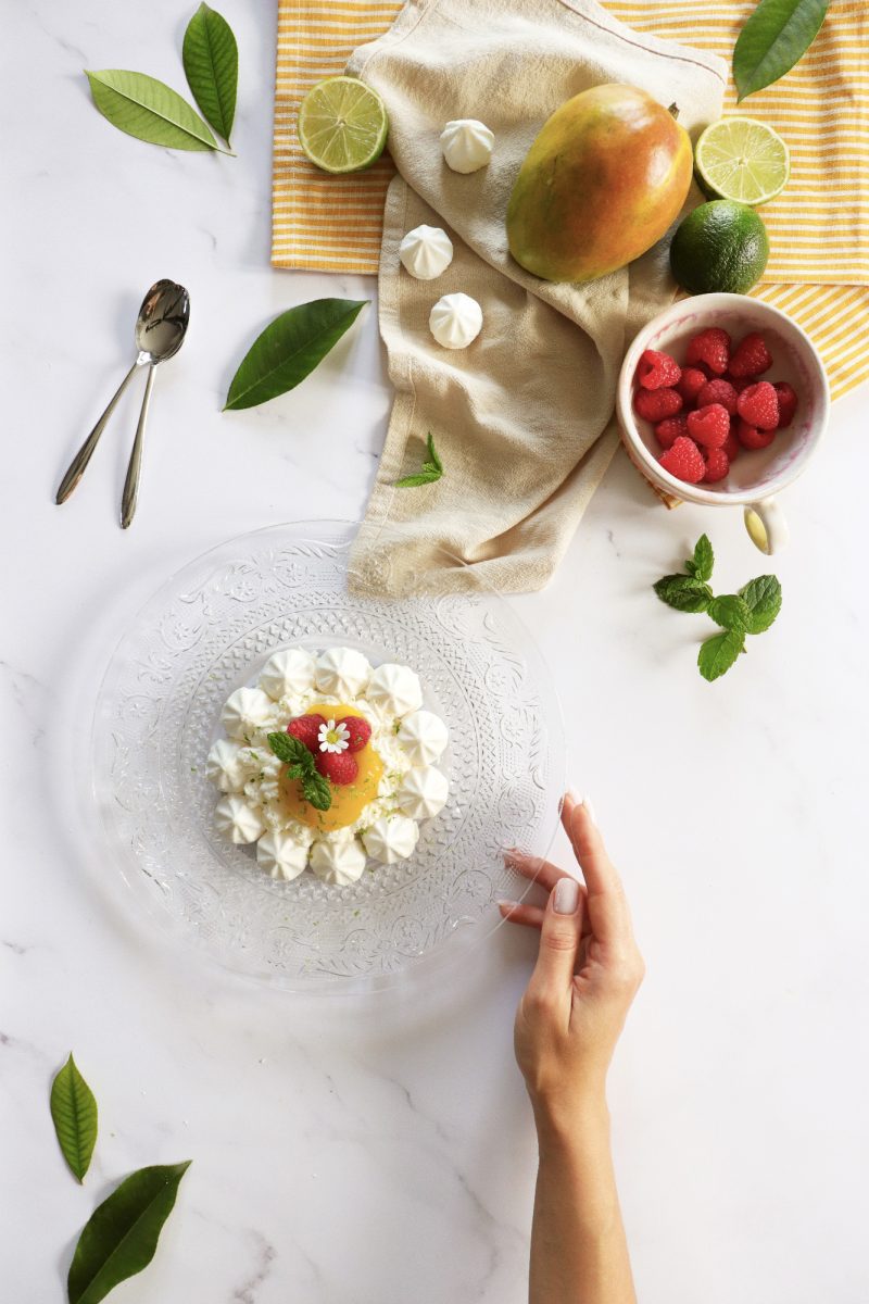 Vista dall’alto di una mini pavlova con salsa al mango e lamponi, circondata da lime, mango, meringhe e una tazza di lamponi, con mano che impugna il piatto.