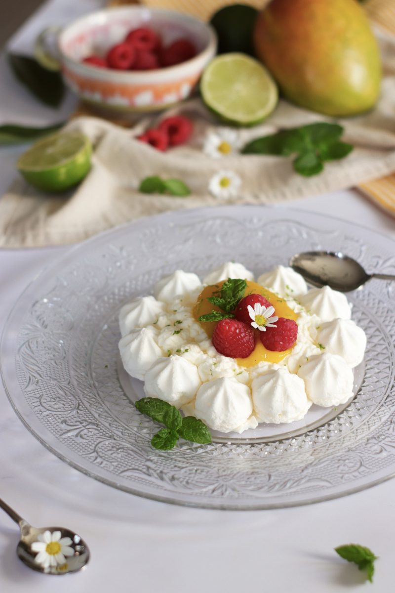 Mini pavlova al mango con panna montata, lamponi freschi e foglie di menta su piatto di vetro decorato, dessert estivo monoporzione.