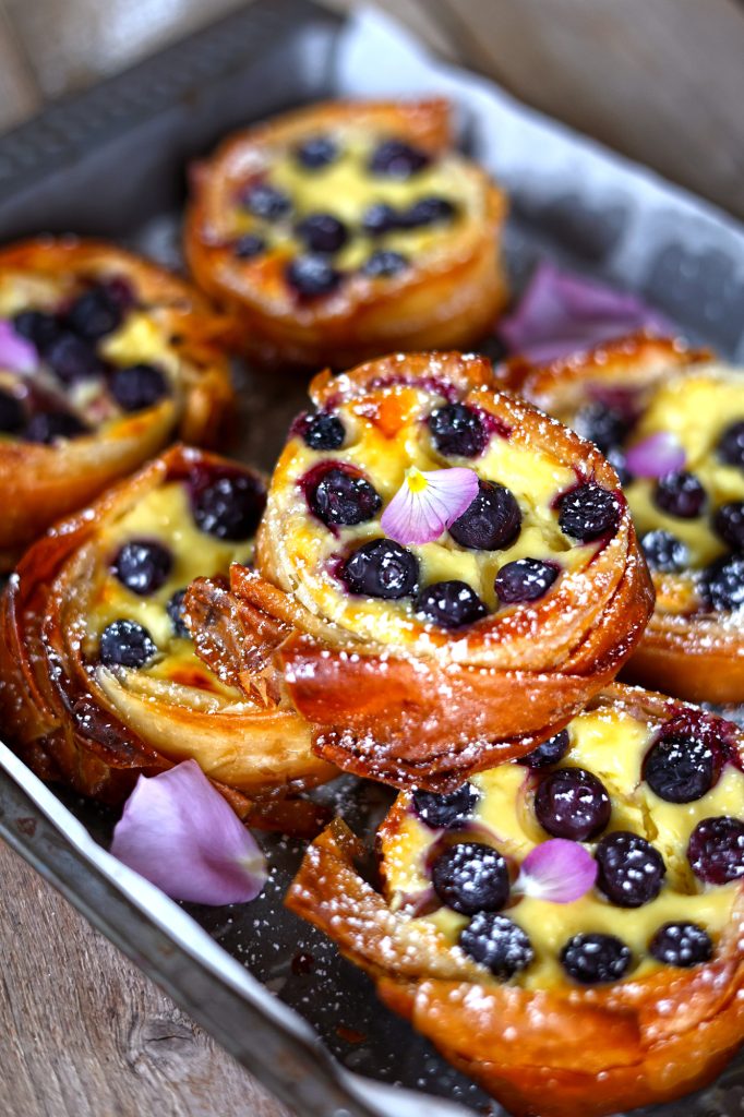 Close-up sulle mini cheesecake tart ai mirtilli e limone in una teglia da forno con qualche petalo di fiore rosa